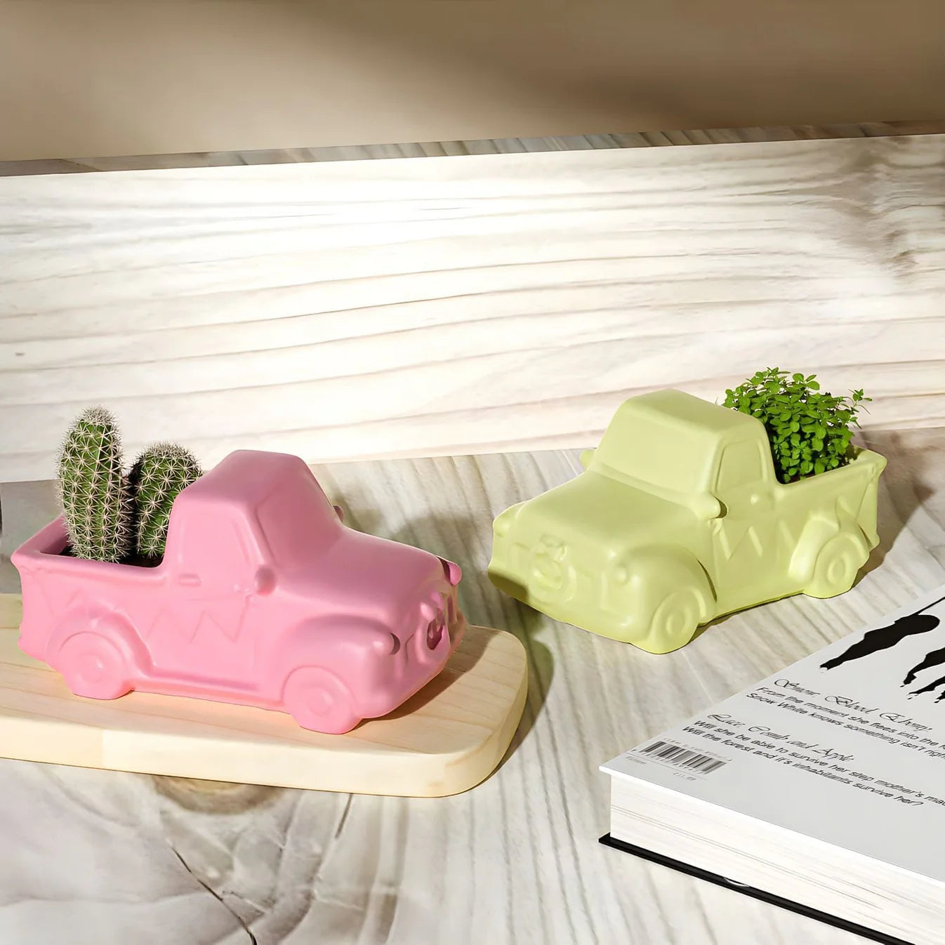 Two truck-shaped planters, one pink and one green, with plants inside on a wooden surface.