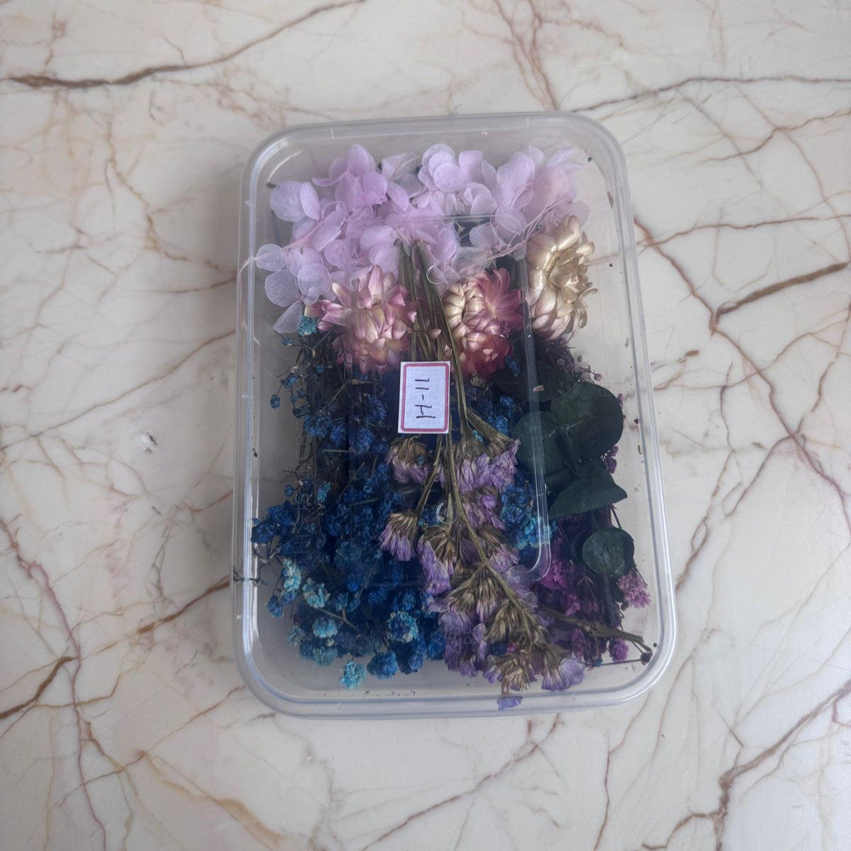 Assorted dried flowers in a clear plastic container on a marble surface